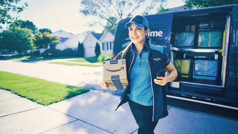 A smiling Amazon delivery driver walks carrying a package to deliver. The delivery vehicle is parked on the street.