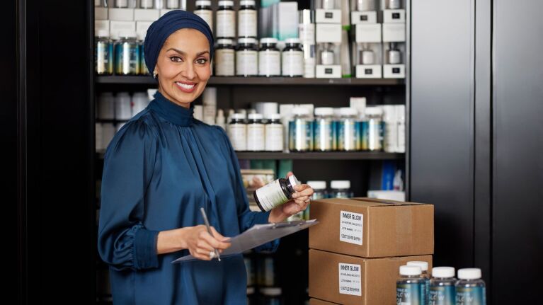 A photo of Dr. Noreen Galaria, co-founder, Inner Glow, holing a container of vitamins and a clipboard. There are shipping boxes behind her.