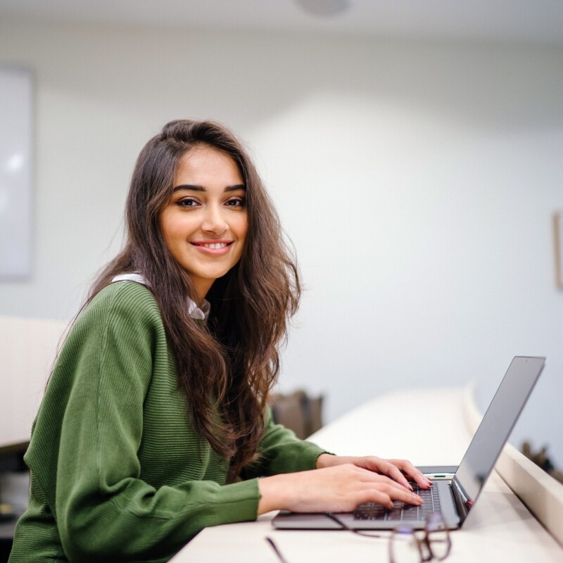 A young woman wearing a green sweatshirt smiles at the camera while studying on her laptop.