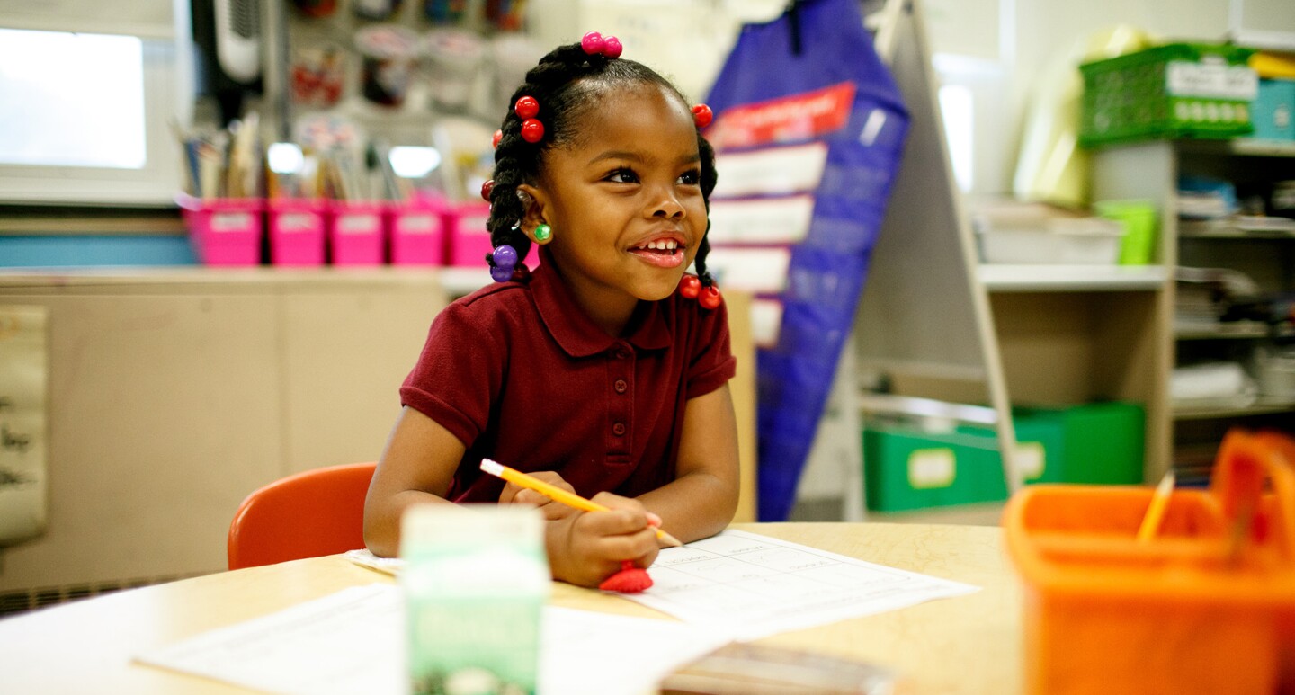 A young girl in a school setting sits with a pen and paper. Blurred out in front of her is a milk carton and snack.