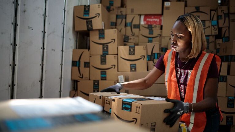 An image of an Amazon employee in an orange work vest lifting a package in a fulfillment center.