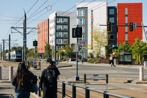 Two people dressed for cool weather walk along a sidewalk. Across the street are a number of condo buildings. A mass transit vehicle approaches the intersection. 