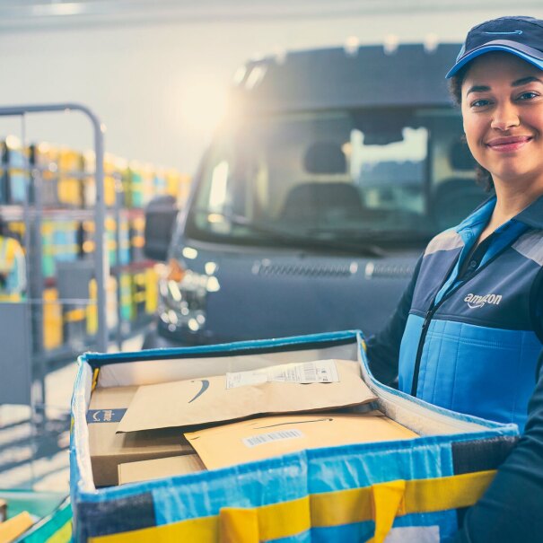 A photo of a delivery driver standing at an Amazon fulfillment center, holding a box that contains Amazon packages. A delivery van is behind them.