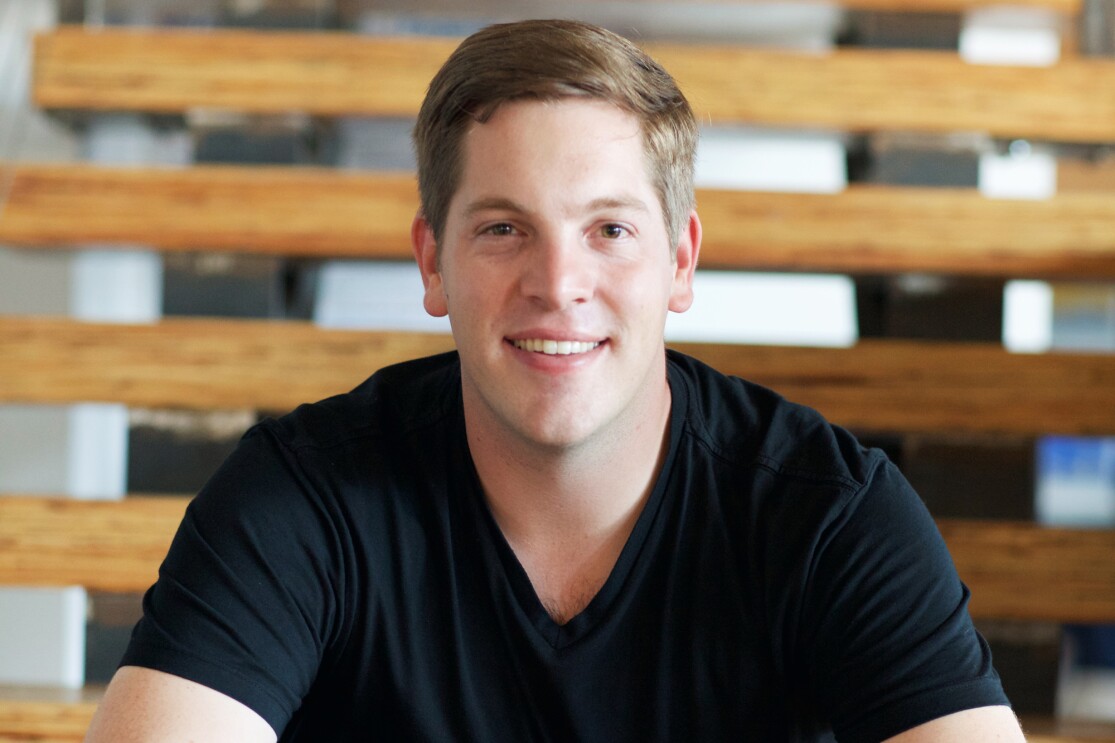 Professional headshot of a man wearing a black v-neck shirt sitting against a wood paneled wall.