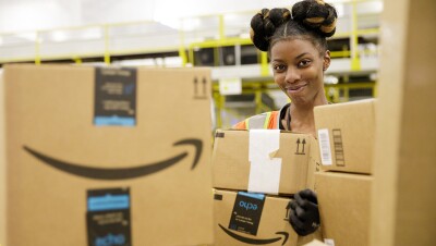 A photo of an employee holding two Amazon Prime delivery boxes. They are standing inside an Amazon fulfillment center.