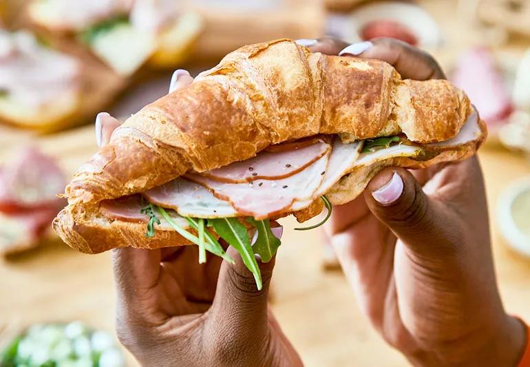 Person making sandwich with deli meat and a croissant with veggies.