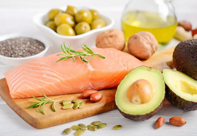 A large piece of salmon on a cutting board surrounded by avocado, olive oil and nuts