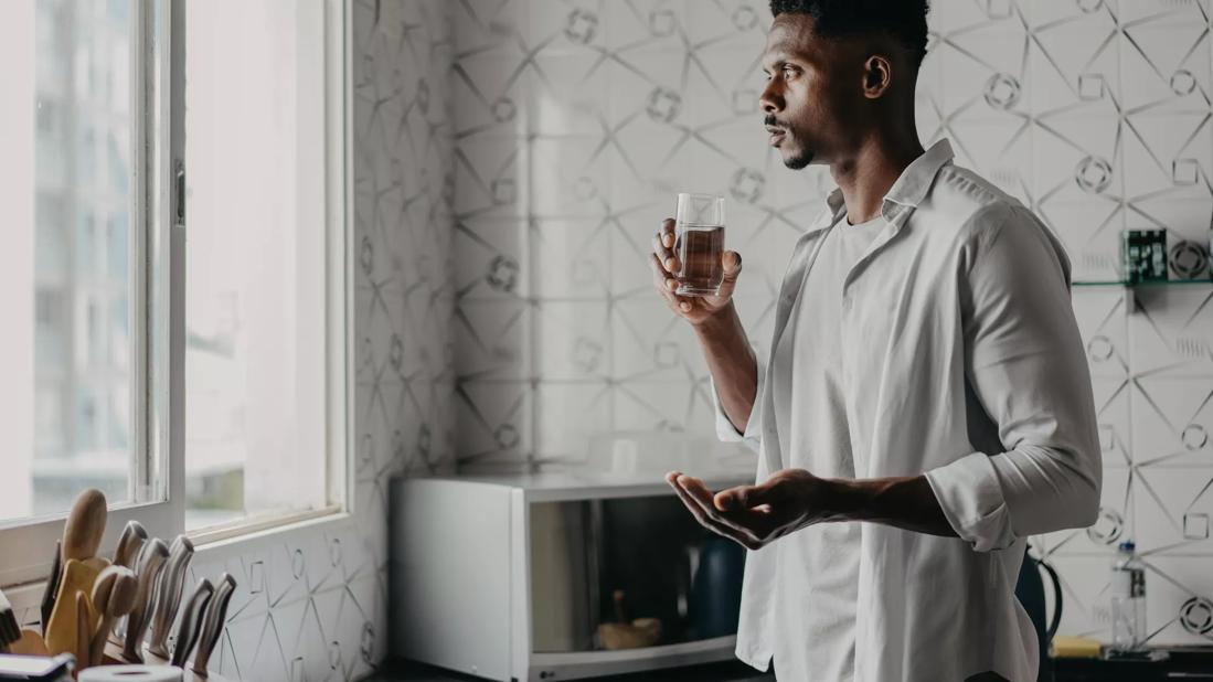 Person standing in kitchen holding glass of water in one hand and medication in the other