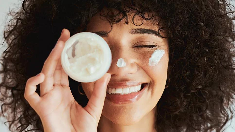 Smiling person holding small container of moisturizer close to face, with product applied to face