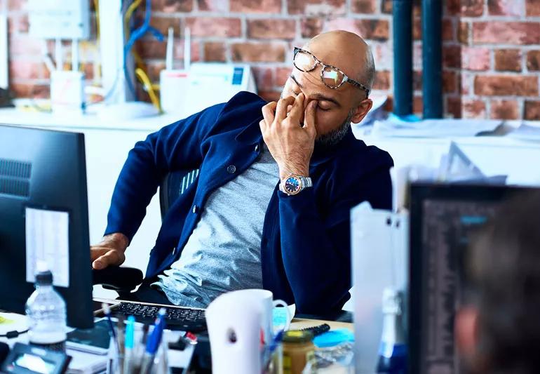 A person sitting at their desk, rubbing their eyes