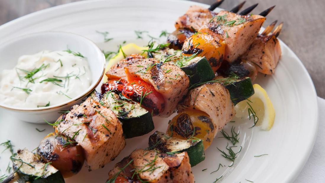 Salmon and vegetable kebabs covered in herbs next to a small dish of white sauce