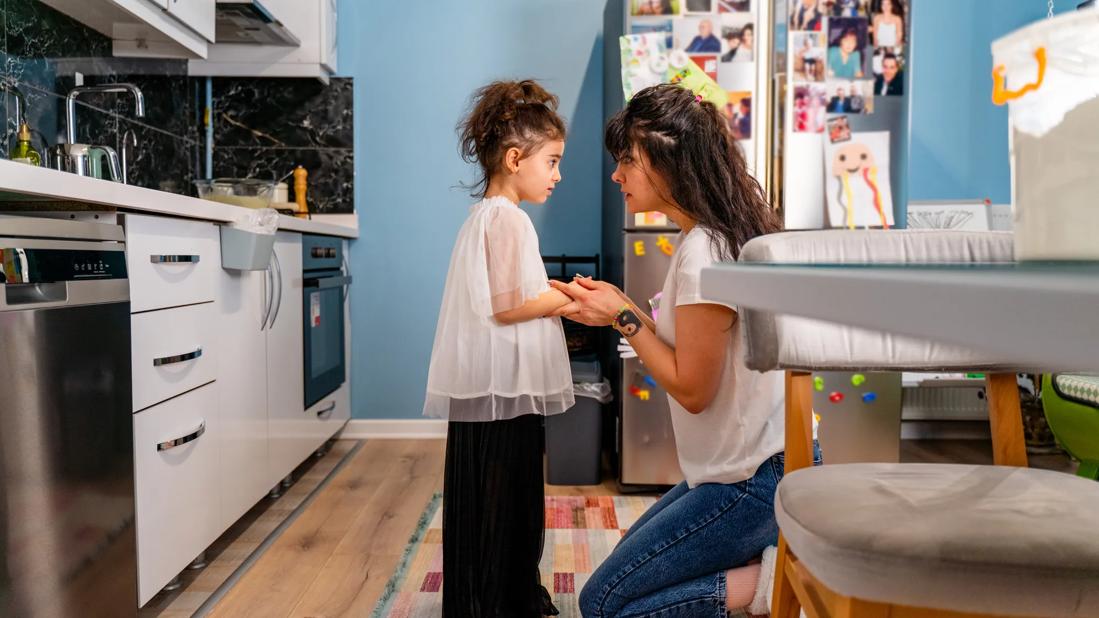 Caregiver kneeling down at home, holding child's hands, with stern look on caregiver's face