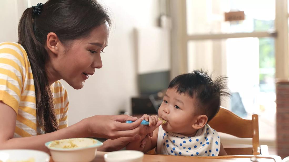 Caregiver spoon feeding baby in highchair at the table