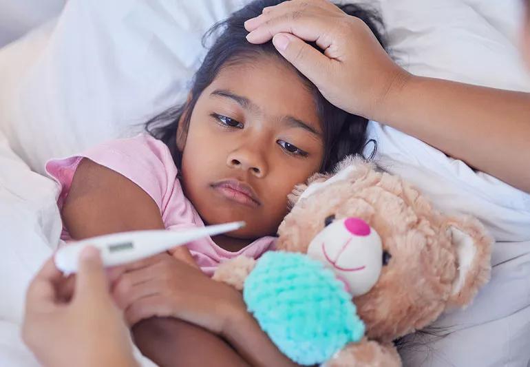 Child in bed sick with parent's hand in foreground holding thermometer.