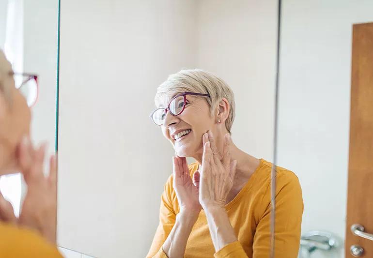 A smiling woman with short gray hair touching her cheeks while looking in the mirror