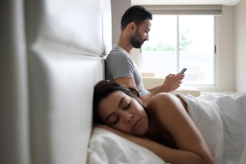 A couple in bed, one sleeping, one on their cellphone, in early morning light