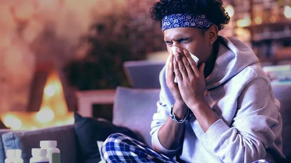 Sick person on couch using tissue on nose with medication bottles on coffee table