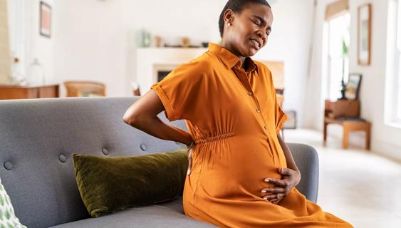 Pregnant woman sitting on couch at home holding her stomach and back, wincing in discomfort