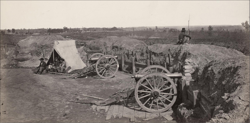 The photograph shows two cannons and a tent behind a barricade of dirt and sandbags. A group of men huddle in the tent. One man sits on top of the barricade holding his bayonet rifle and looks out at the wasteland in front of him.