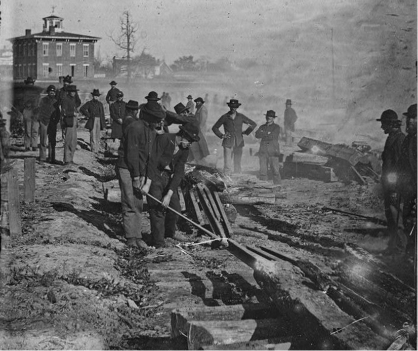 A group of men stand around a railroad that has only the cross ties remaining. Two men work to break the track.