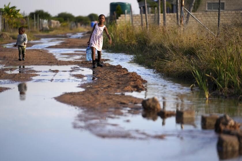 A young girl with a water bottle and her brother walk through a flooded street caused by an overflowing reservoir in Hamanskraal, Pretoria, South Africa, Friday, May 26, 2023. Health authorities are yet to confirm the exact source of the cholera outbreak, but poor waste water management and local government instability in South Africa's capital city have been blamed for the situation. (AP Photo/Themba Hadebe)