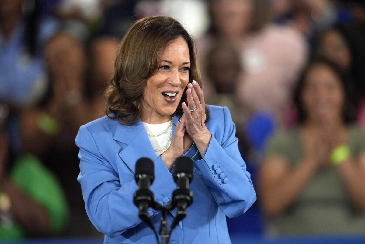 Kamala Harris clapping as she stands in front of a double microphone, the crowd behind her out of focus