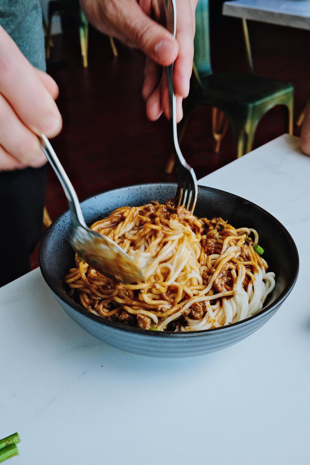 Two hands hold serving utensils that toss dan dan noodles in a black bowl on white tabletop at Highland Park's Mala Class