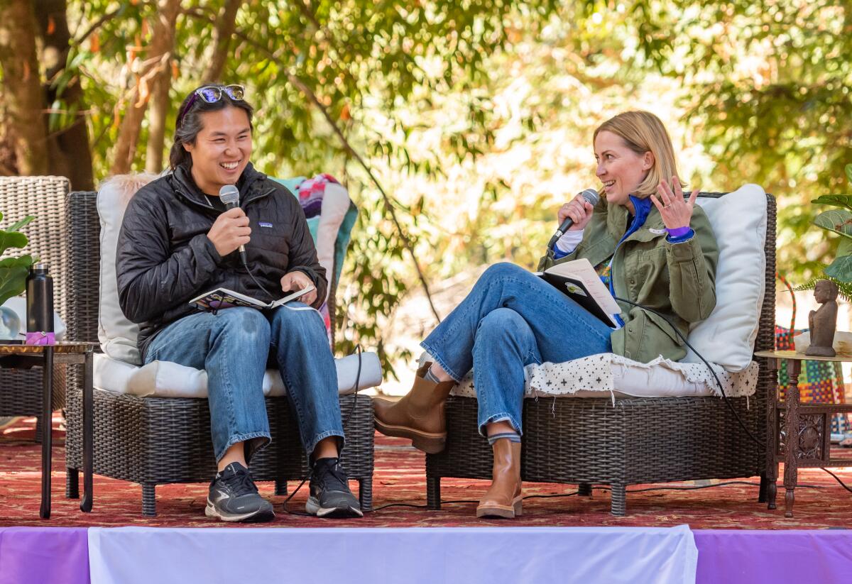 Two people sit in chairs on a stage outdoors while holding microphones.