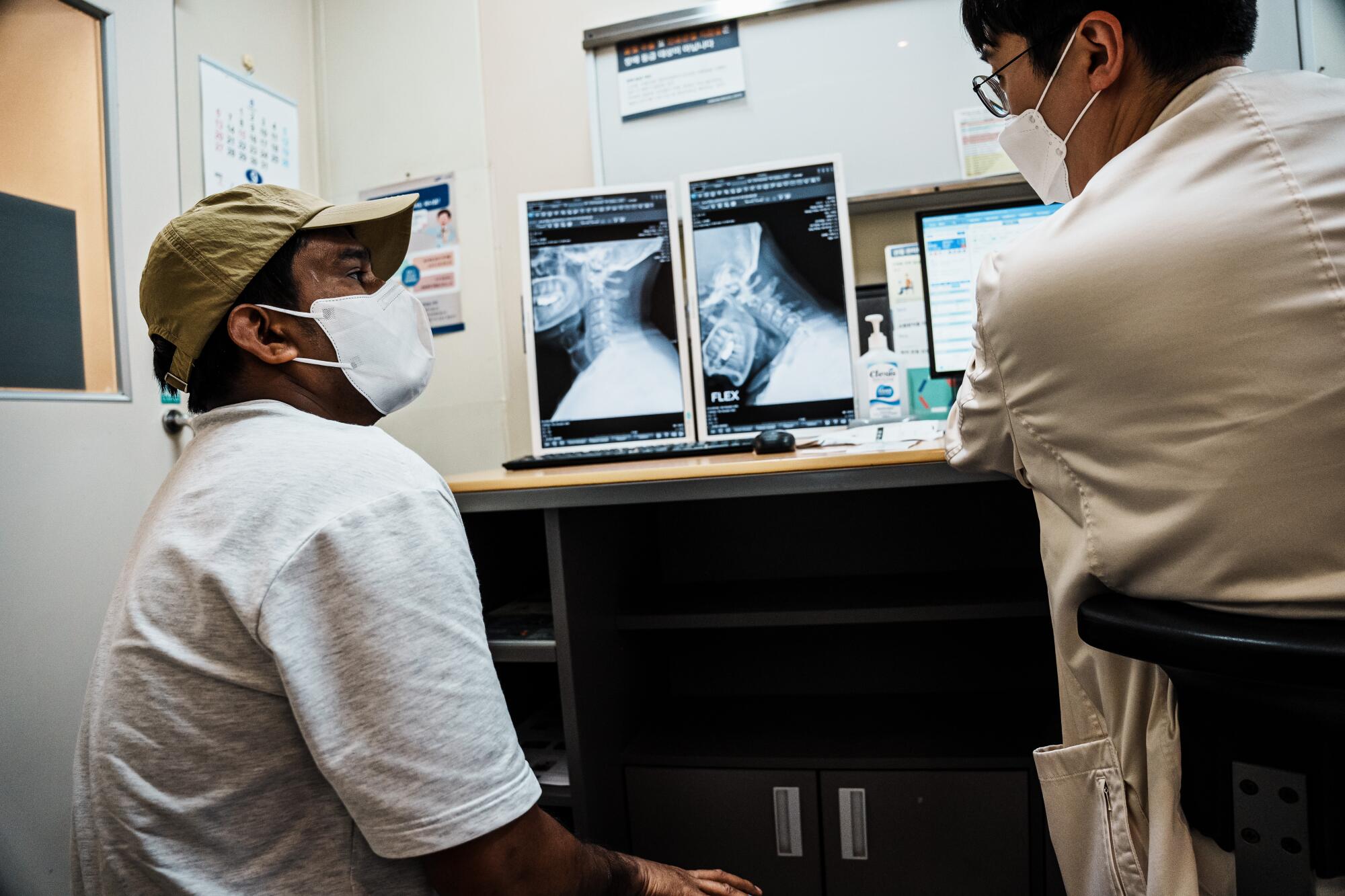 A man in a hat sits looking toward a doctor in a white coat in a medical exam room