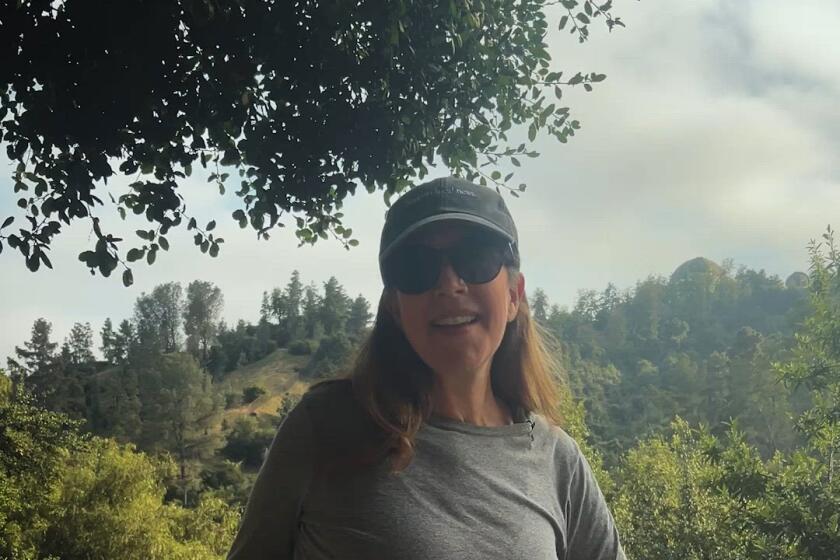 Woman wearing hiking gear stands and stretches in front of a scenic view and greenery