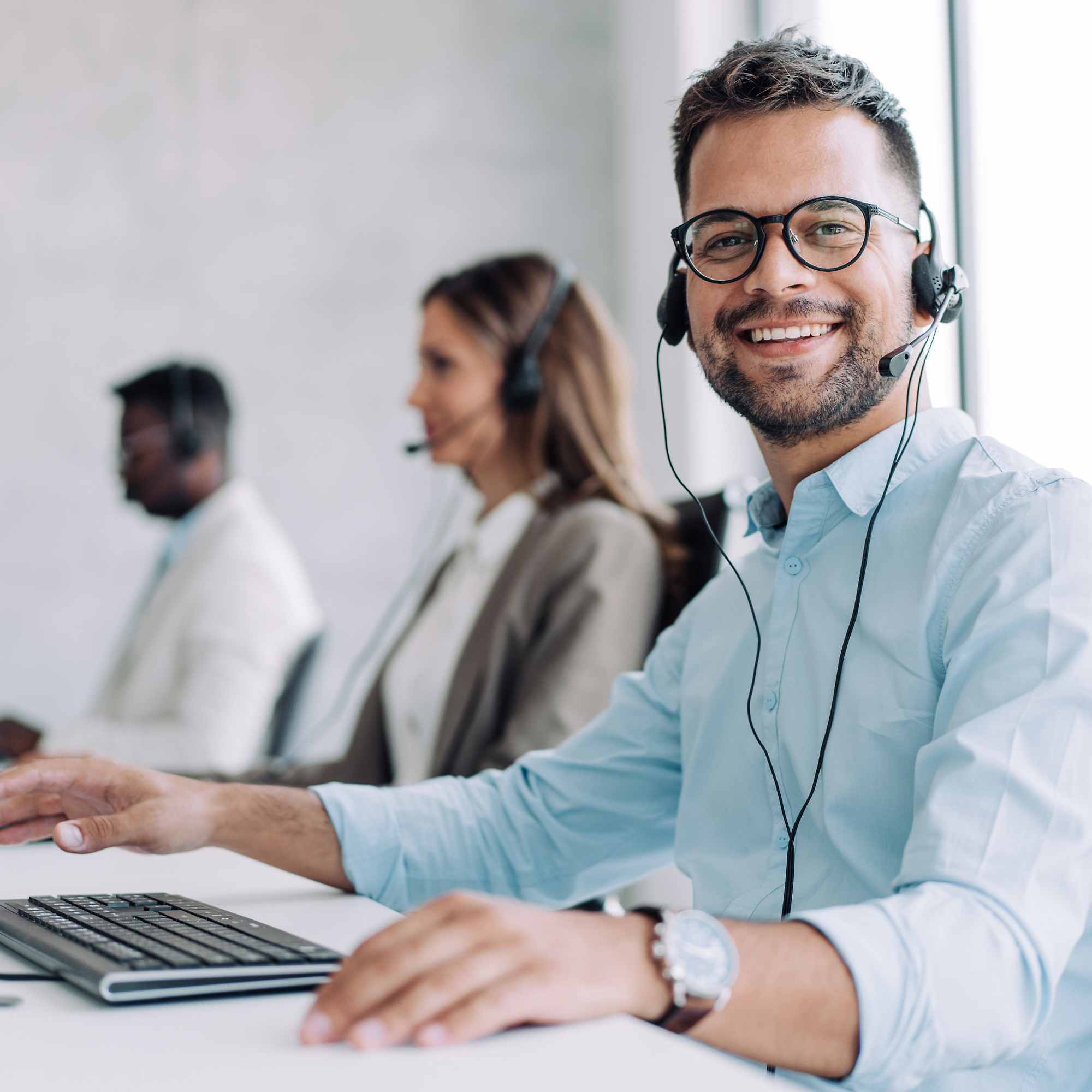 Un hombre que sonríe y lleva gafas con auriculares, trabajando en un escritorio junto a compañeros en una oficina moderna.