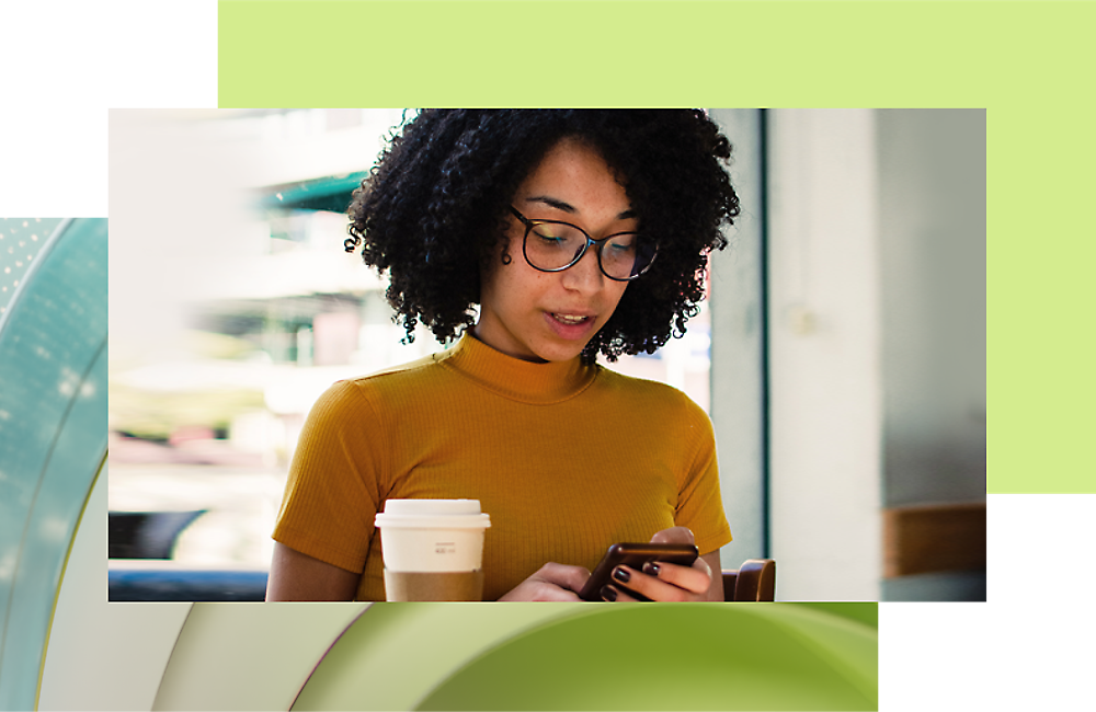 Una mujer con una parte superior amarilla y gafas usando su smartphone mientras sosteniendo una taza de café en un café.