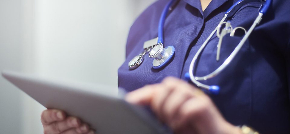 Doctor con un uniforme médico azul sosteniendo una tableta, con un estetoscopio alrededor del cuello, revisando los registros médicos.