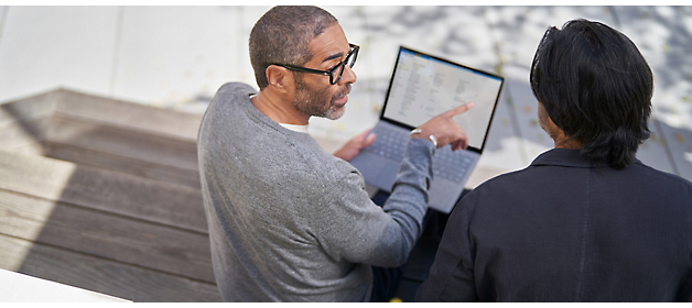 Dos hombres examinando la pantalla de un portátil juntos al aire libre, uno apuntando a la pantalla mientras participan en una discusión.