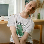 Portrait of a man in a chair, looking towards the camera. He wears a t-shirt with a cat dressed up as a dinosaur.