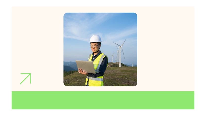 A man wearing a reflective vest and hardhat stands outside looking at a laptop with a mountainous landscape filled with wind turbines in the background.