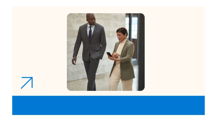 Two colleagues dressed in formal business attire walking together in a hallway looking at a mobile device.