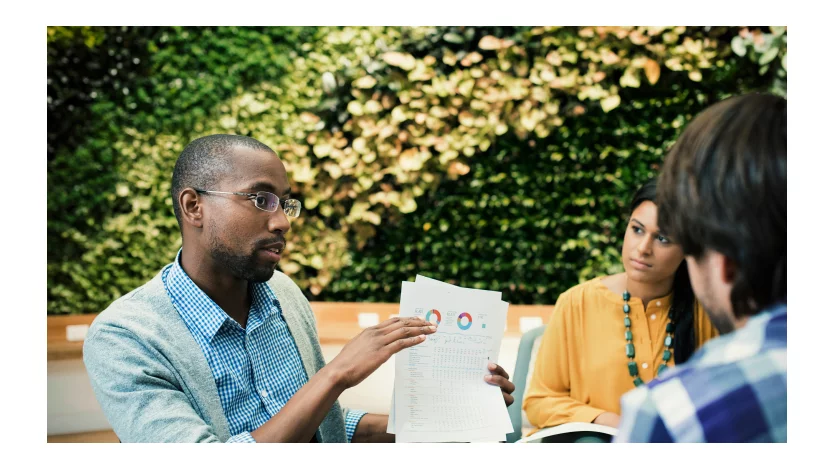 A man holds a document containing charts and discusses them with two business colleagues.