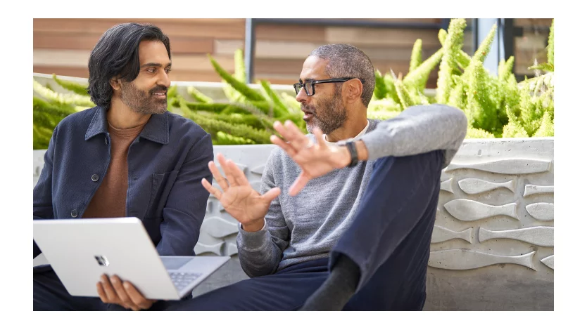 Two men have a discussion while one holds a laptop. 