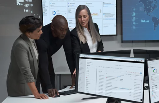 Three technology workers looking at a computer screen in an office environment with surrounding monitor screens displaying reports and maps.