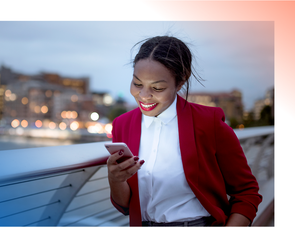 Una mujer sonriente con una chaqueta roja usando un smartphone en un puente de ciudad al atardecer.