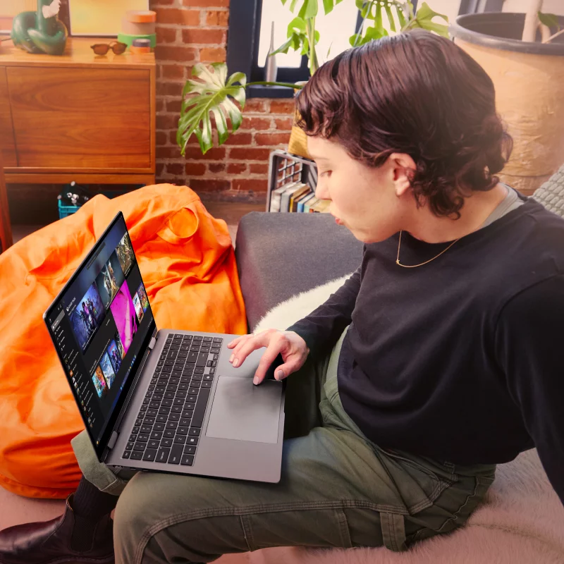 A person sitting on a couch looking at PC games on an open laptop with a bean bag, amp, shelf, and potted plant in the background.