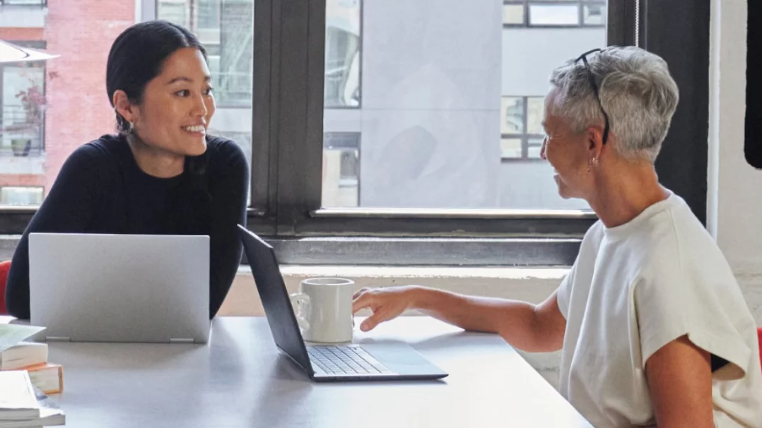 Deux femmes discutent à un bureau, des ordinateurs portables Windows ouverts face à elles.