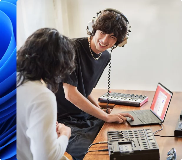 Un jeune homme souriant porte un casque audio. Une autre personne compose de la musique sur un ordinateur portable avec un clavier électronique.