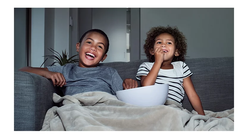 Two children share a bowl of snacks and a blanket while enjoying a movie at home.