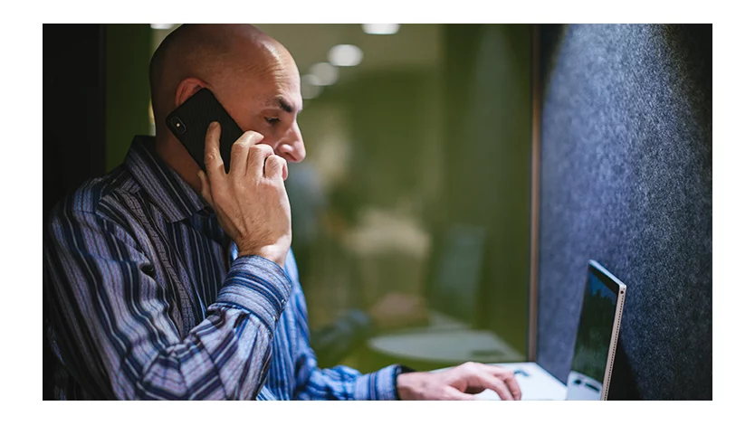 A man talks on a cell phone while working on a computer in an isolated booth.