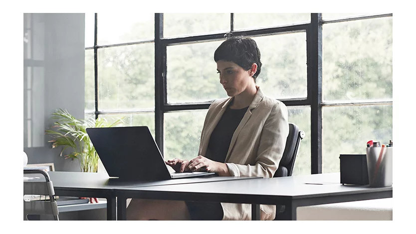 A woman is sitting at a long desk and typing on a laptop in a modern window office that has glass walls.