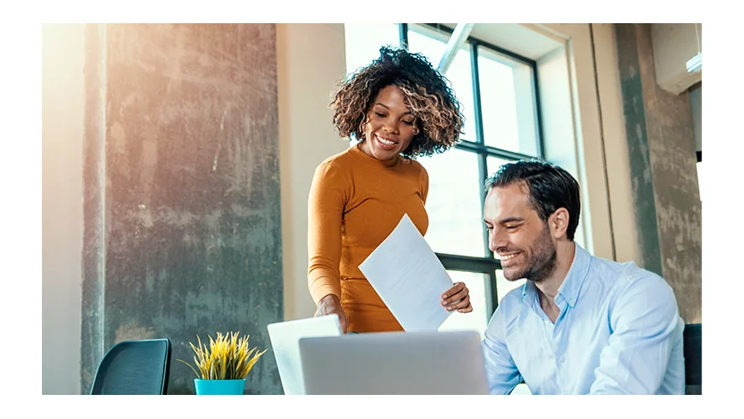 Woman standing in an office looks at a laptop screen with a seated male colleague.