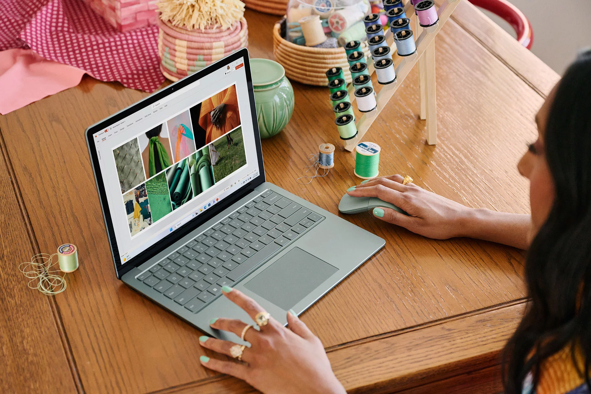 A young woman sits at a desk filled with multiple spools of thread, fabric, and other sewing materials while interacting with her Surface device.