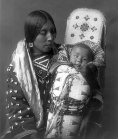 A Crow woman holding an infant in a decorated cradleboard, photograph by Edward S. Curtis, c. 1908.